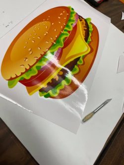 A glossy print of a burger illustration with layers of lettuce, tomato, onion, cheese, patty, and bun, placed on a table next to a cutting tool.