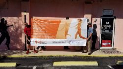 Two men hold a banner promoting body contouring and SculpSure services at Indigo Dermatology, with a contact number (321) 951-1010. The banner is displayed in front of a pink wall.