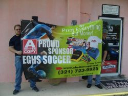 Two individuals hold a large banner in front of a store entrance that reads "A Better Copy Proud Sponsor EGHS Soccer" and includes the company's contact information and promotional images.
