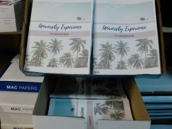 Image shows several stacks of "University Experience FYE 1000 Workbook" books with palm tree covers in an open cardboard box. The box is on a shelf with other boxes labeled "MAC Papers.