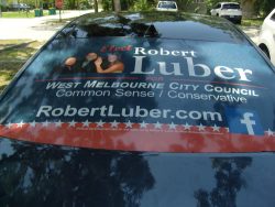 Rear window of a car displaying a campaign ad for Robert Luber, running for West Melbourne City Council, featuring a website and a Facebook icon. The ad promotes common sense and conservative values.